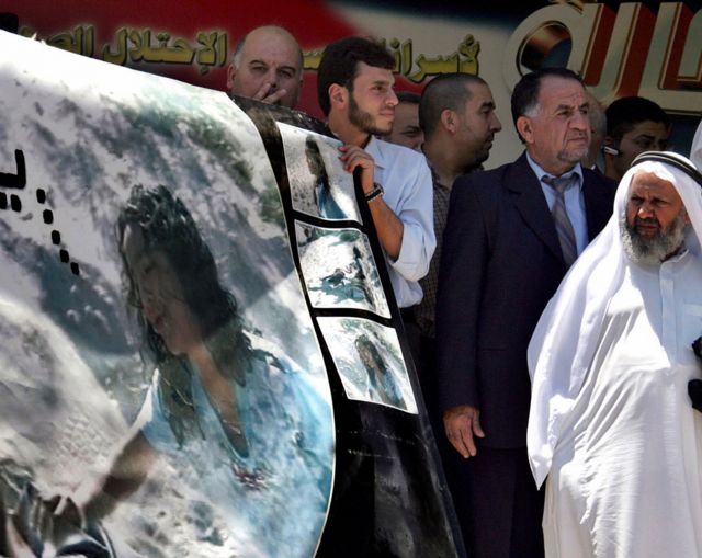 A pooster of Huda Ghalia reaching out to her fatyer's body while protesters hold the poster up