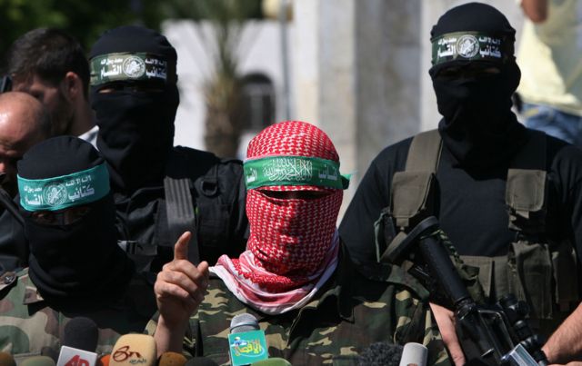 Abu Ubaida standing at a microphone podium with two masked gunmen standing dehind him