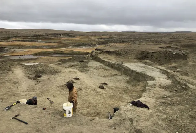 A wide view shows a barren brown landscape, with two people in the foreground on a dig with tools around them, on a cloudy grey day.